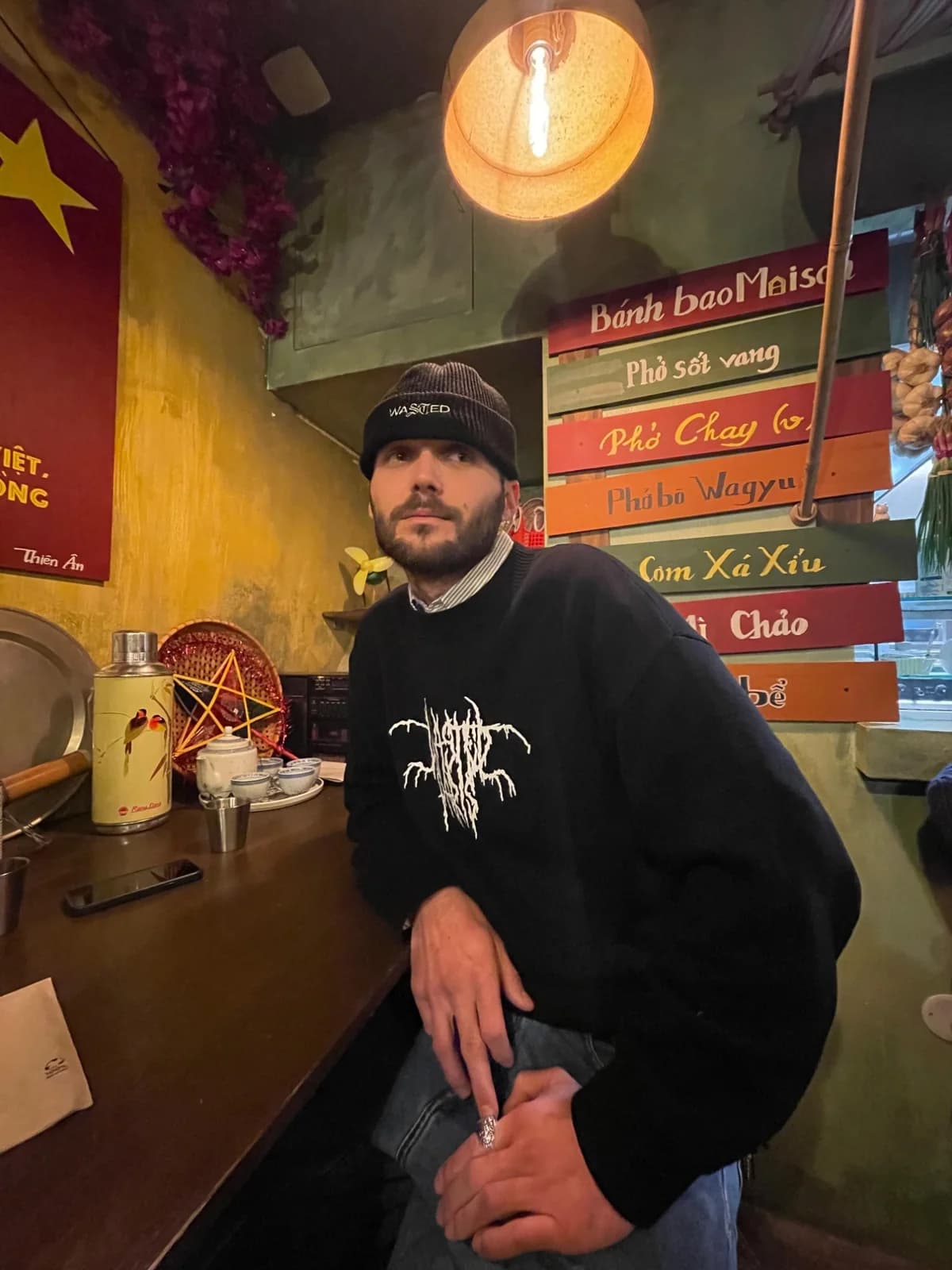 Portrait de Hugo Bona, en bonnet et pull noirs, accoudé au comptoir d’un restaurant aux murs colorés.