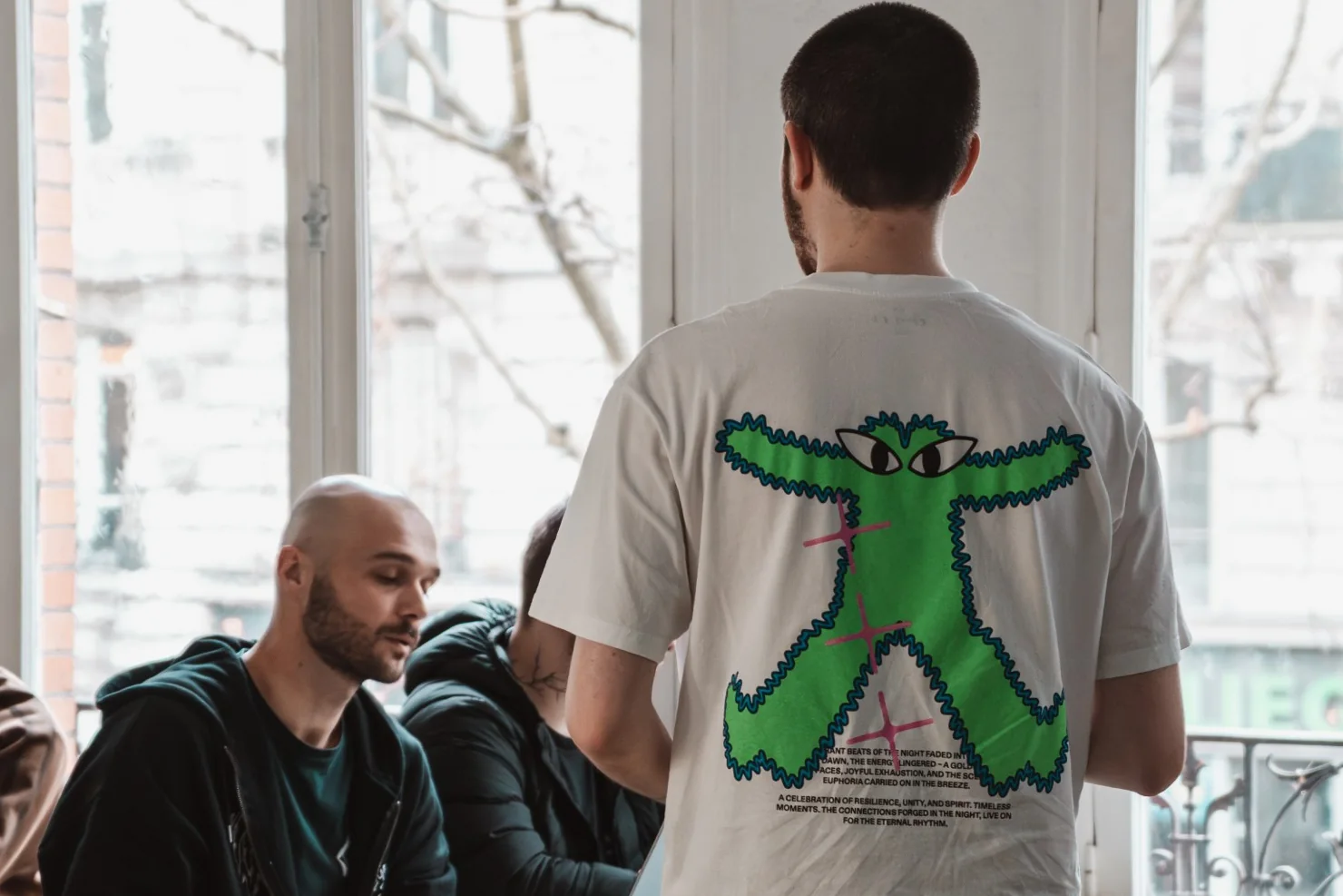 Séance de travail en classe à l’ESD, devant de grandes fenêtres : un étudiant assis à son ordinateur échange avec un camarade debout.